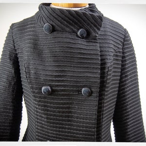 May include: A black ribbed jacket with a high collar and four black buttons. The jacket has a double-breasted closure.