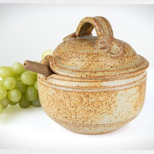 Peut inclure: Un pot à miel en céramique avec un couvercle et un égouttoir en bois. Le pot est de couleur brun et beige moucheté. Le couvercle a une poignée et un motif décoratif.