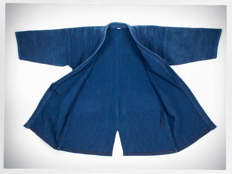 May include: A dark blue denim jacket with a traditional Japanese style, open and laid flat. The jacket is made of a thick, textured fabric and has a simple design.