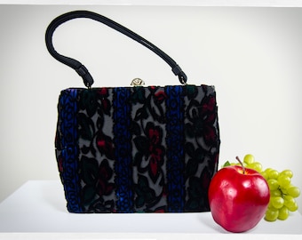 May include: A vintage handbag with a black handle and a floral pattern in shades of blue, red, and green. The bag has a clasp with a decorative jewel. A red apple and green grapes are next to the bag.