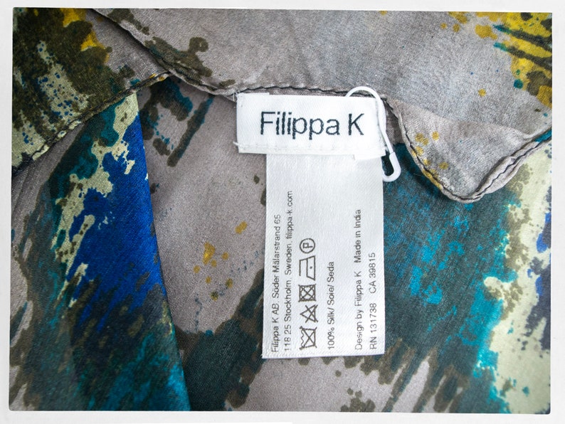 May include: A white tag with black text that reads "Filippa K" and includes care instructions and company information. The tag is attached to a gray and blue patterned fabric.