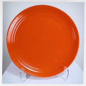 May include: A bright orange ceramic plate with concentric ring detailing. The plate is round and has a glossy finish. The plate is displayed on a clear acrylic stand, showcasing its bright colour and design.