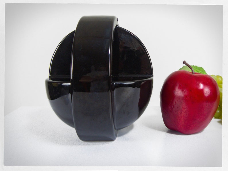 K&ouml;nnte beinhalten: Eine schwarze Keramikschale mit einem einzigartigen Design, geformt wie eine Kugel mit einem Ring um die Mitte. Die Schale steht auf einer wei&szlig;en Oberfl&auml;che neben einem roten Apfel.