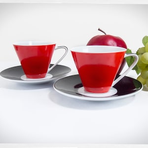 May include: Two red and white ceramic coffee cups with black and white saucers. The cups are cone-shaped and have a white rim. The saucers have a black center and a white rim.