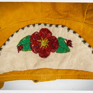 May include: A close-up of a beaded flower design on a piece of yellow leather. The flower is red with green leaves and white beads around the edge.
