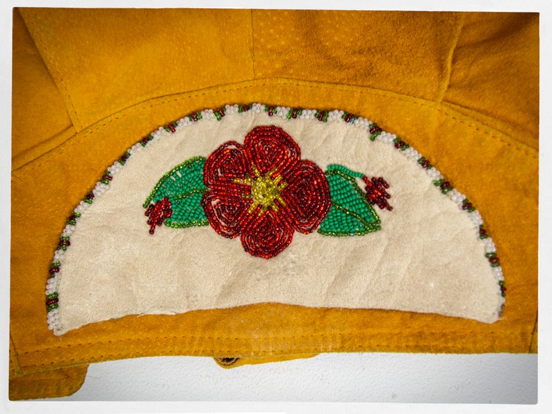 May include: A close-up of a beaded flower design on a piece of yellow leather. The flower is red with green leaves and white beads around the edge.