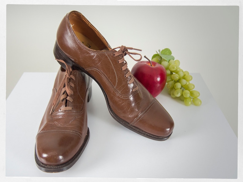May include: Two brown leather lace-up shoes with a white background. A red apple and a bunch of green grapes are in the background.