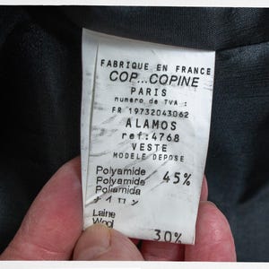 May include: A close-up of a clothing label with text in French, including "FABRIQUE EN FRANCE" and "COP...COPINE PARIS". The label also lists "ALAMOS" and "VESTE" with percentages of Polyamide and Wool.