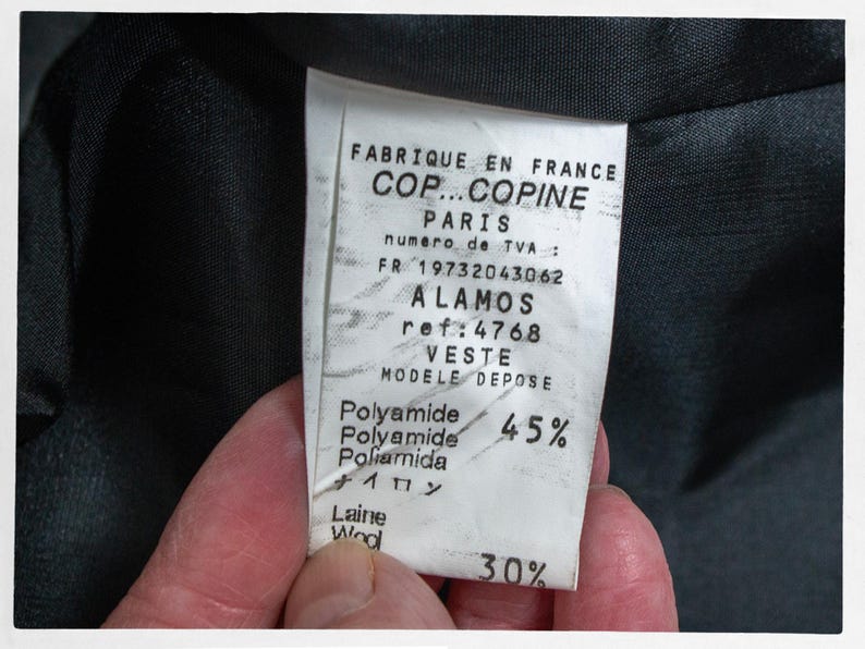 May include: A close-up of a clothing label with text in French, including "FABRIQUE EN FRANCE" and "COP...COPINE PARIS". The label also lists "ALAMOS" and "VESTE" with percentages of Polyamide and Wool.