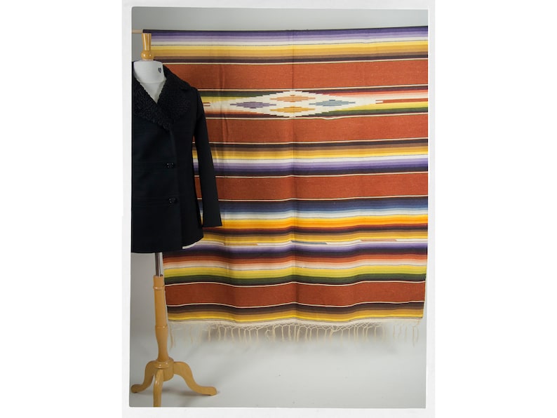 May include: A colorful striped woven blanket with a diamond pattern in the center. The blanket is displayed on a wooden stand.