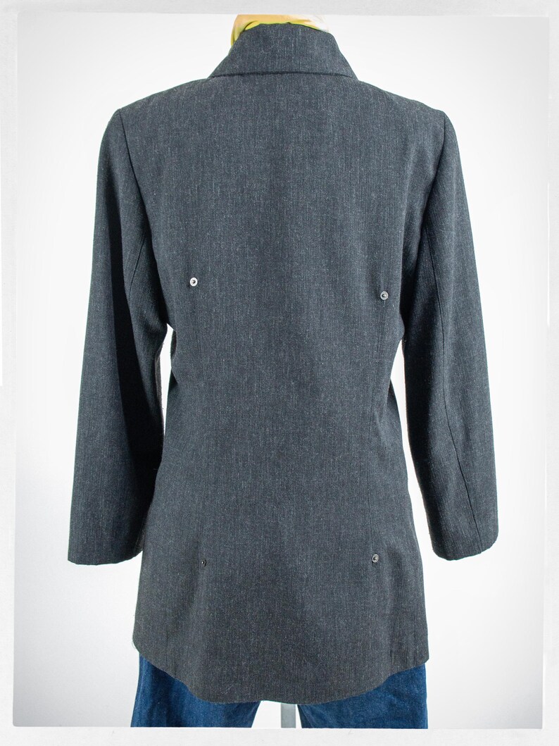 May include: A dark gray blazer with long sleeves, viewed from the back. The blazer has a notched collar and three small buttons on the back. The blazer is displayed on a mannequin, with blue jeans visible at the bottom.