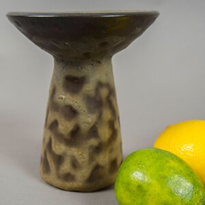 May include: A brown ceramic pedestal with a wide, shallow top and a textured, mottled surface. The pedestal is sitting on a gray surface with two lemons beside it.