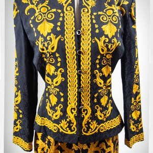 Puede incluir: Una chaqueta y falda de traje de seda con estampado negro y dorado. La chaqueta tiene un cuello mandar&iacute;n y cierre de botones en la parte delantera. La falda es de l&iacute;nea A y tiene una cintura alta.