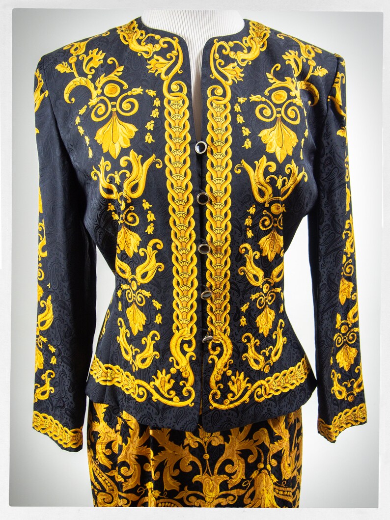 Puede incluir: Una chaqueta y falda de traje de seda con estampado negro y dorado. La chaqueta tiene un cuello mandar&iacute;n y cierre de botones en la parte delantera. La falda es de l&iacute;nea A y tiene una cintura alta.