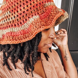 Op de afbeelding: Een handgemaakte gehaakte bucket hat in tinten oranje, rood en bruin. De hoed heeft een textuur, open geweven ontwerp en een licht naar beneden hellende rand. De hoed is een stijlvol accessoire voor bescherming tegen de zon.