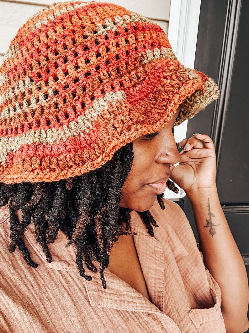 Op de afbeelding: Een handgemaakte gehaakte bucket hat in tinten oranje, rood en bruin. De hoed heeft een textuur, open geweven ontwerp en een licht naar beneden hellende rand. De hoed is een stijlvol accessoire voor bescherming tegen de zon.