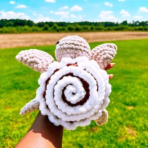 Puede incluir: Un peluche de tortuga hecho a mano con ganchillo y un diseño de caparazón en espiral. La tortuga tiene una cabeza y extremidades de color marrón claro, con un caparazón en espiral blanco y marrón. El peluche se sostiene sobre un fondo de hierba verde y cielo azul.