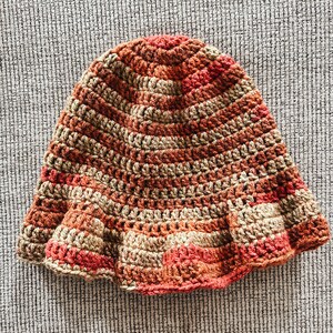 Op de afbeelding: Een handgemaakte gehaakte hoed in tinten bruin, beige en rood. De hoed heeft een uitlopende rand en een gestructureerd steekpatroon. De hoed is een warm getint accessoire.