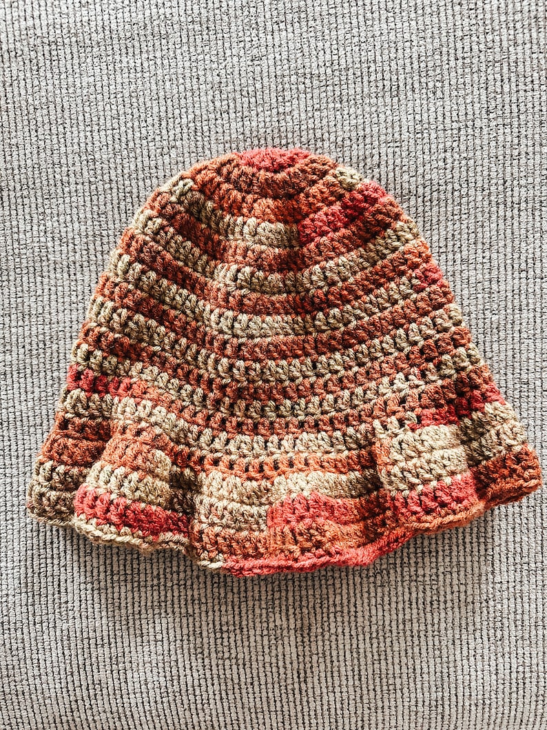 Op de afbeelding: Een handgemaakte gehaakte hoed in tinten bruin, beige en rood. De hoed heeft een uitlopende rand en een gestructureerd steekpatroon. De hoed is een warm getint accessoire.