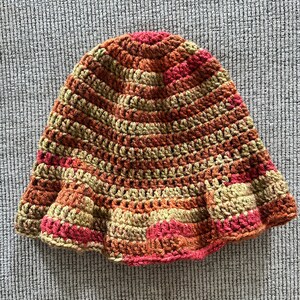 Op de afbeelding: Een handgehaakte hoed in warme tinten rood, oranje en beige. De hoed heeft een uitlopende rand en een gestreept patroon. Deze stijlvolle hoed is perfect om een vleugje handgemaakte charme aan elke outfit toe te voegen.