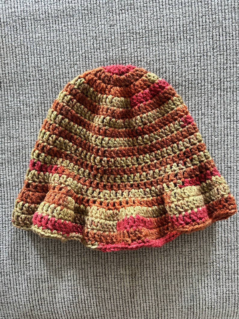 Op de afbeelding: Een handgehaakte hoed in warme tinten rood, oranje en beige. De hoed heeft een uitlopende rand en een gestreept patroon. Deze stijlvolle hoed is perfect om een vleugje handgemaakte charme aan elke outfit toe te voegen.