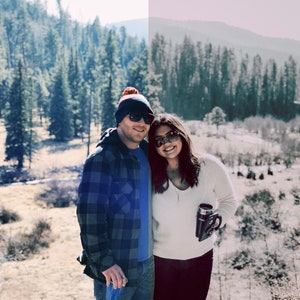 May include: A couple wearing winter clothing poses in front of a snowy forest. The man is wearing a blue and black plaid shirt, a blue beanie, and sunglasses. The woman is wearing a white sweater, black pants, and sunglasses. She is holding a black travel mug.