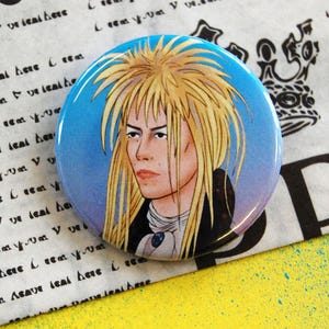 May include: A circular badge with a portrait of a person with long, blonde hair against a blue and purple gradient. The badge has a shiny finish and is set against a patterned paper background with black text and a yellow and teal surface.