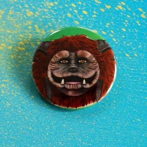 May include: A circular badge with a cartoon monster design. The creature has brown fur, a large smile, and visible teeth. The badge has a green top and is set against a blue background with yellow paint splatters.