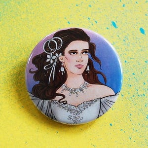 May include: A circular badge with a portrait of a woman with brown hair, wearing a white dress and a jeweled necklace. The background fades from purple to blue. The badge is set against a yellow backdrop.