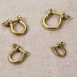 May include: Four gold-coloured metal D-rings of varying sizes. The D-rings are on a beige fabric background.
