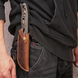 May include: A wood-handled knife with a patterned blade is secured in a brown leather sheath. The sheath is attached to a person's belt, with a brass-colored snap closure. The knife and sheath are set against a dark background.