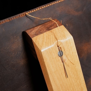 May include: Close-up of a leather item with a wooden component. The leather is dark brown with a textured appearance. The wooden piece is light-coloured with a darker wood accent. A needle and thread are attached.