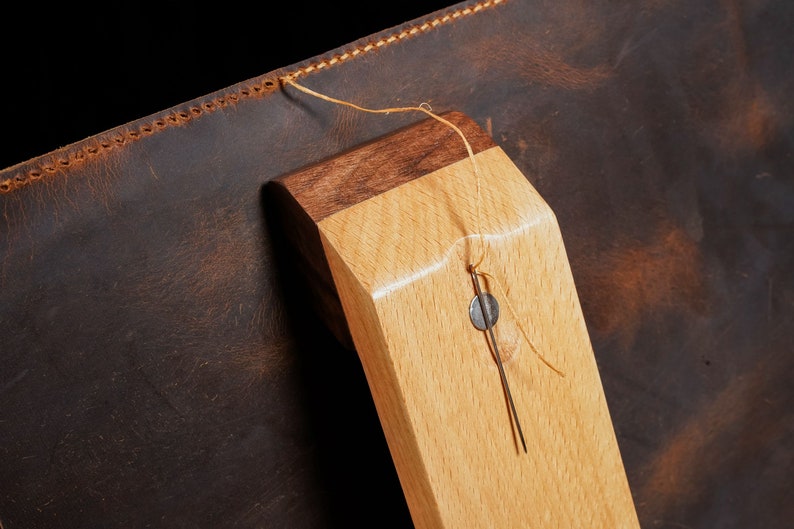 May include: Close-up of a leather item with a wooden component. The leather is dark brown with a textured appearance. The wooden piece is light-coloured with a darker wood accent. A needle and thread are attached.