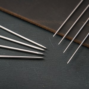 May include: A set of six silver stitching awls, also known as leather needles, are arranged in a fan shape on a dark gray surface. The awls are made of metal and have a sharp point at one end. They are used for hand-stitching leather.