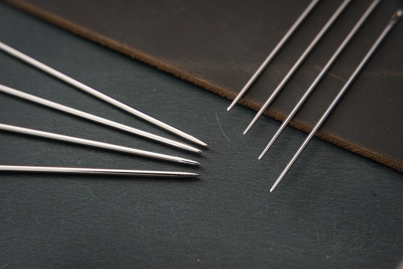 May include: A set of six silver stitching awls, also known as leather needles, are arranged in a fan shape on a dark gray surface. The awls are made of metal and have a sharp point at one end. They are used for hand-stitching leather.
