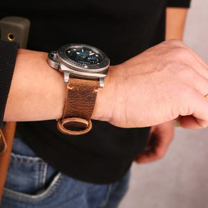 May include: A stainless steel wristwatch with a dark blue face and a brown leather strap. The watch has a large, round face with luminous hands and markers. The strap is made of distressed leather and has a buckle closure. The watch is worn on a wrist.