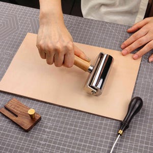 May include: A leather craft setup featuring a wooden-handled roller, a wooden edge creaser, and a leather working knife. The roller is chrome with a wooden handle. The leather is a light tan color, and the tools are on a gray cutting mat.