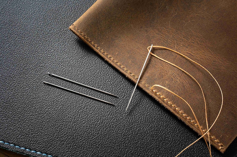 May include: Two silver needles and a length of brown waxed thread on a dark brown leather surface. The thread is being used to stitch the leather together.