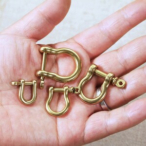 May include: Four gold-coloured metal D-rings of varying sizes. The D-rings are arranged in a hand.