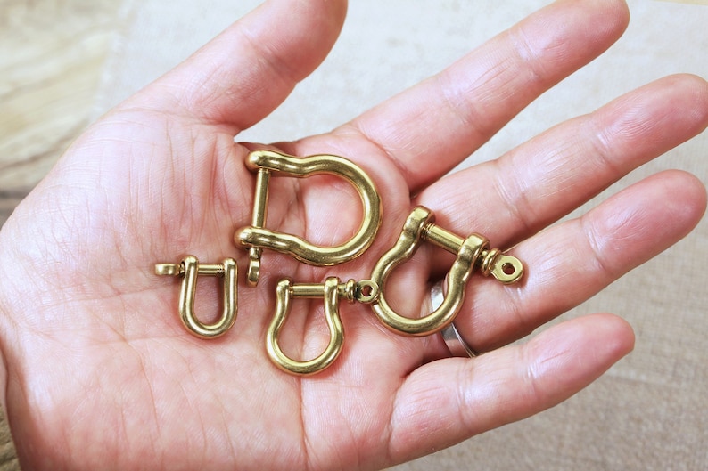 May include: Four gold-coloured metal D-rings of varying sizes. The D-rings are arranged in a hand.