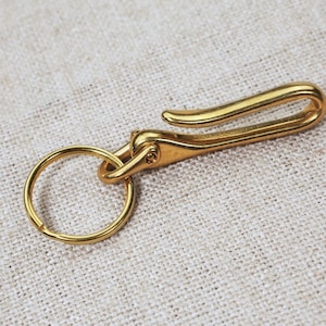 Könnte beinhalten: Ein goldfarbener Schlüsselanhänger aus Metall mit einem Haken und einem Ring. Der Haken ist gebogen und der Ring ist rund.