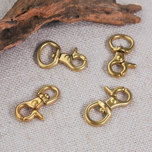 May include: Four gold-toned metal swivel clasps, also known as trigger snaps, are arranged on a light beige fabric surface. The clasps are commonly used for attaching straps, leashes, or other accessories.