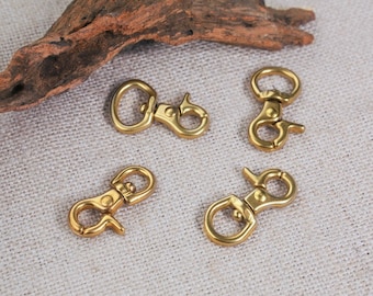 May include: Four gold-toned metal swivel clasps, also known as trigger snaps, are arranged on a light beige fabric surface. The clasps are commonly used for attaching straps, leashes, or other accessories.