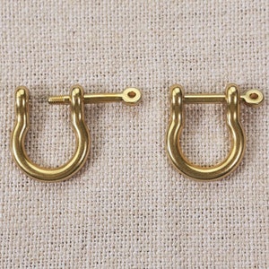 May include: Two gold-toned metal D-rings with a screw-on clasp. The D-rings are on a beige fabric background.