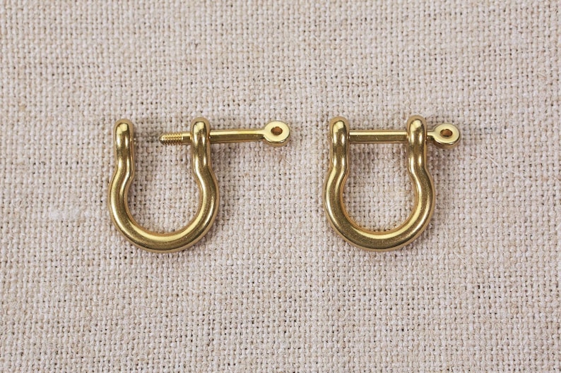 May include: Two gold-toned metal D-rings with a screw-on clasp. The D-rings are on a beige fabric background.