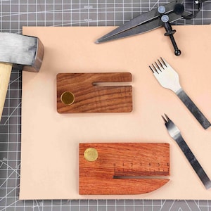 May include: Leathercraft tools including a hammer, dividers, and stitching chisels, arranged on a light brown leather surface. Two wooden measuring tools with brass accents are also present, showcasing a craft-focused setup.