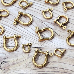 May include: A collection of gold-toned metal u-shaped clasps or hooks. The clasps are arranged on a light wood surface.