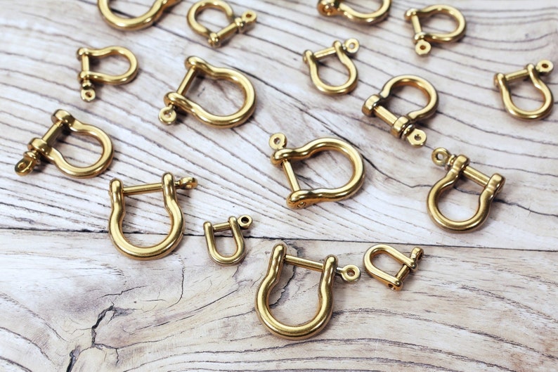 May include: A collection of gold-toned metal u-shaped clasps or hooks. The clasps are arranged on a light wood surface.