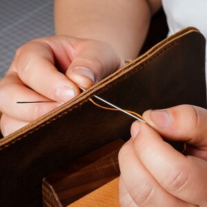 May include: Close-up of a person's hands stitching two pieces of brown leather together with orange thread. The leather is being held in place by a wooden clamp.