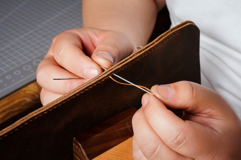 May include: Close-up of a person's hands stitching two pieces of brown leather together with orange thread. The leather is being held in place by a wooden clamp.
